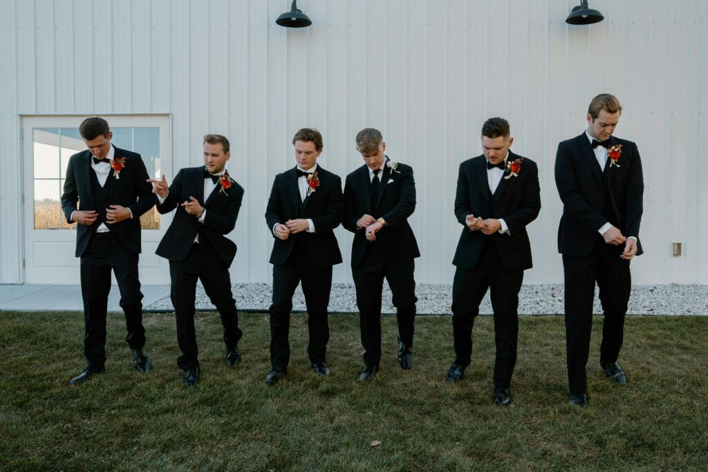Group of groomsmen in tuxedos adjusting cuffs before outdoor wedding ceremony.